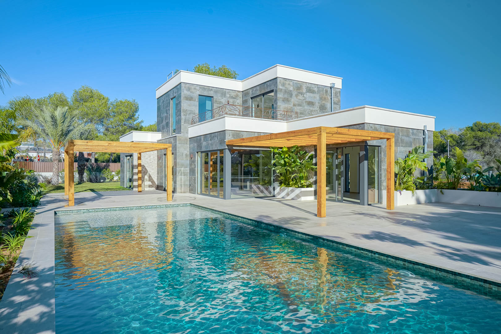 Exterior view featuring a Bali-style pool, spacious tiled terraces, and iroko wood pergolas against a modern facade finished with natural stone cladding.