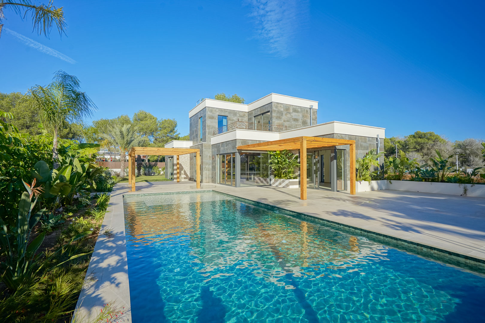Exterior view featuring a Bali-style salt-water pool, large tiled terraces, and iroko wood pergolas against the stone-clad modern facade.