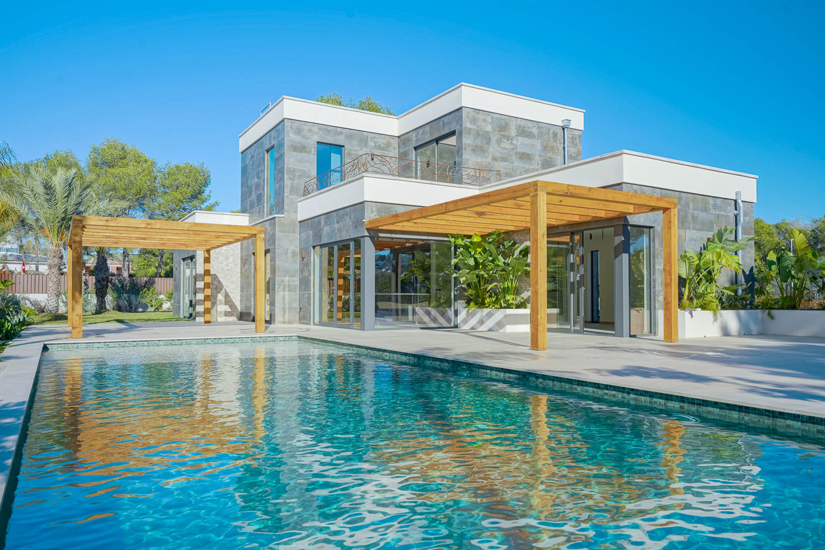 Exterior view of the villa featuring a turquoise mosaic pool, iroko wood pergolas, and a natural stone facade with large glass doors.