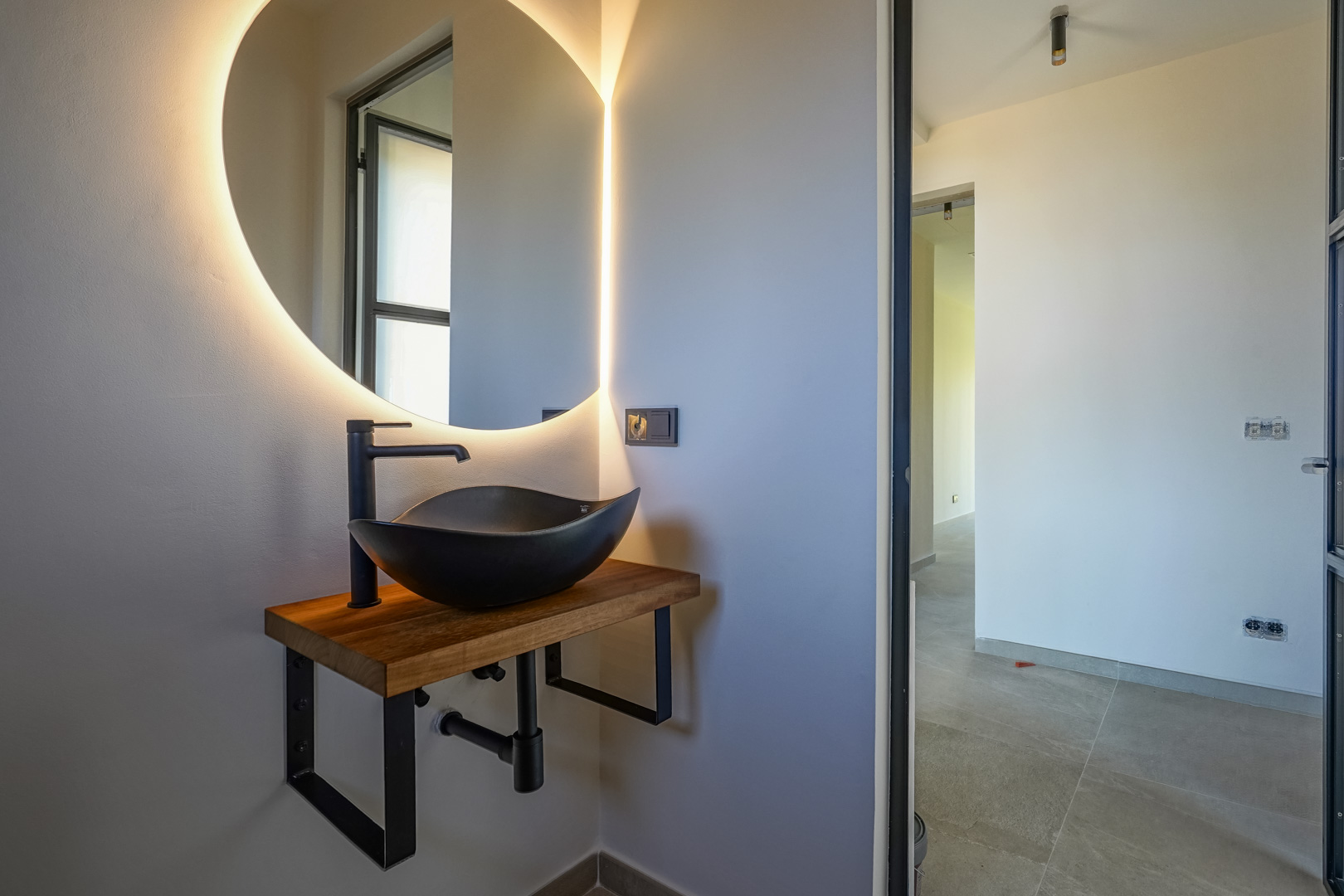 Guest toilet featuring a black vessel sink on a solid wood countertop, complemented by a backlit circular mirror and matte black fixtures.