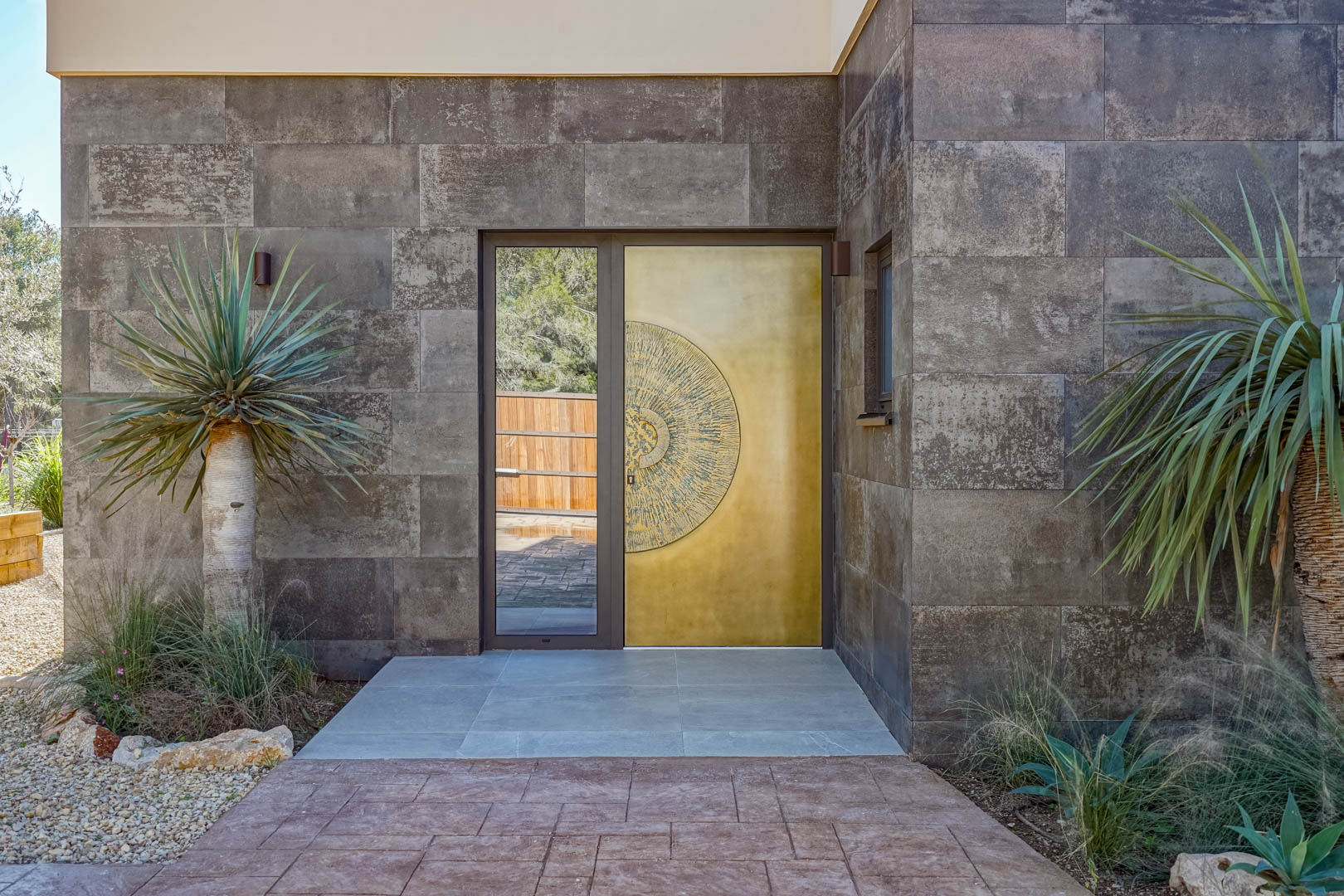 Main entrance featuring a designer golden door with a circular relief, flanked by textured stone walls and architectural landscaping.