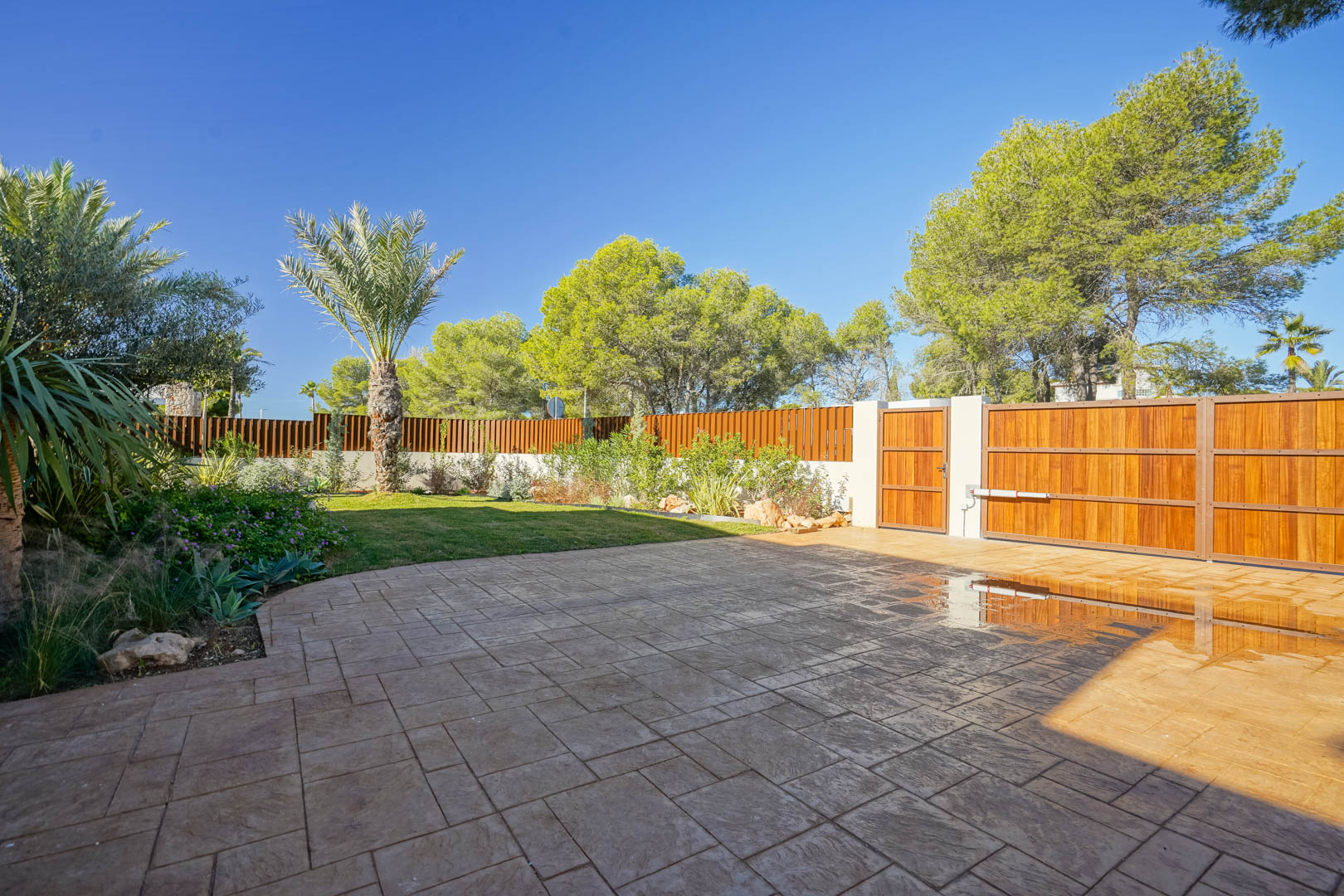 Main entrance featuring a motorized wooden gate, stamped concrete driveway, and a landscaped Mediterranean garden with palm trees and privacy fencing.