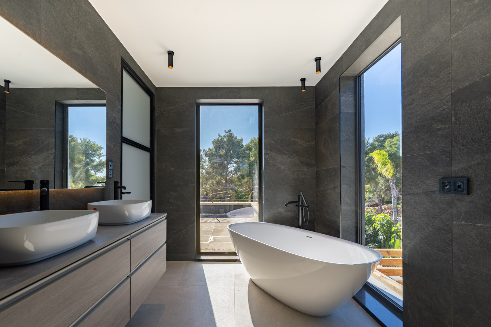 Master bathroom featuring a freestanding oval tub, double vanity with vessel sinks, and floor-to-ceiling windows. Dark stone walls and black fixtures provide a modern contrast.