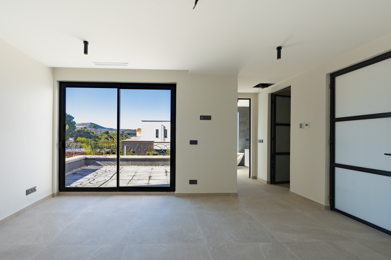 Master bedroom with direct access to a private terrace, porcelain tile floors, and black minimalist lighting. The en-suite bathroom with a tub is visible.