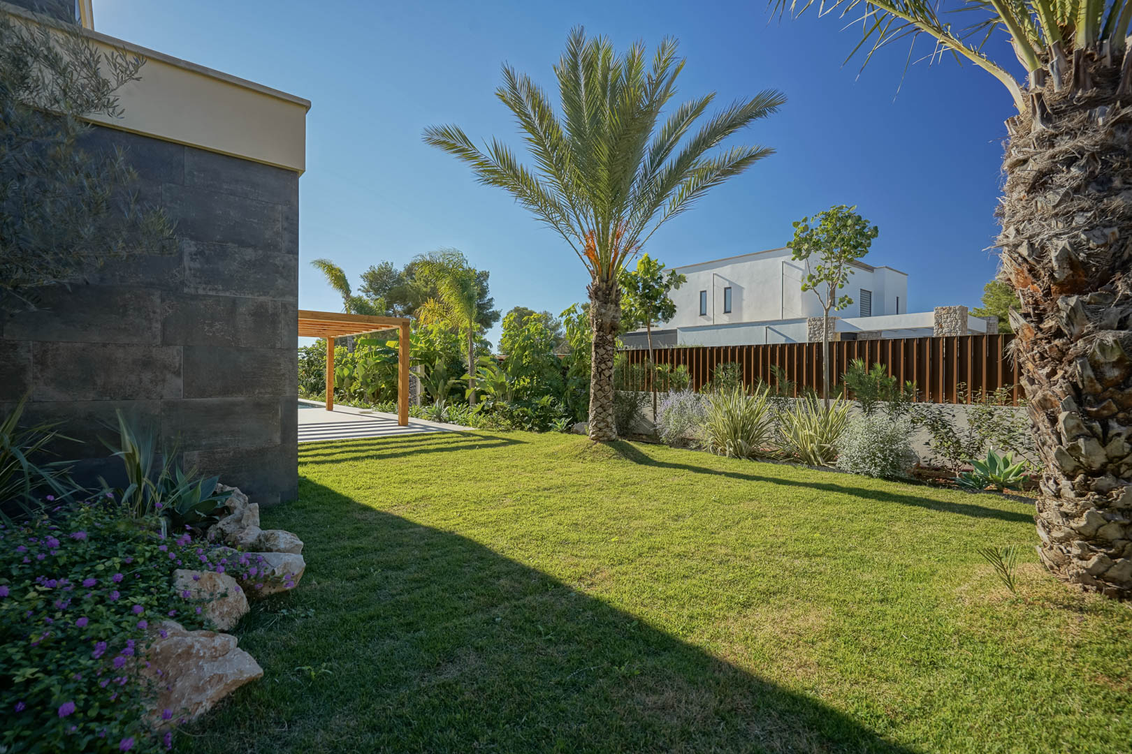 Mediterranean garden featuring a natural lawn, palm trees, and native plants alongside a dark stone facade and an iroko wood pergola.