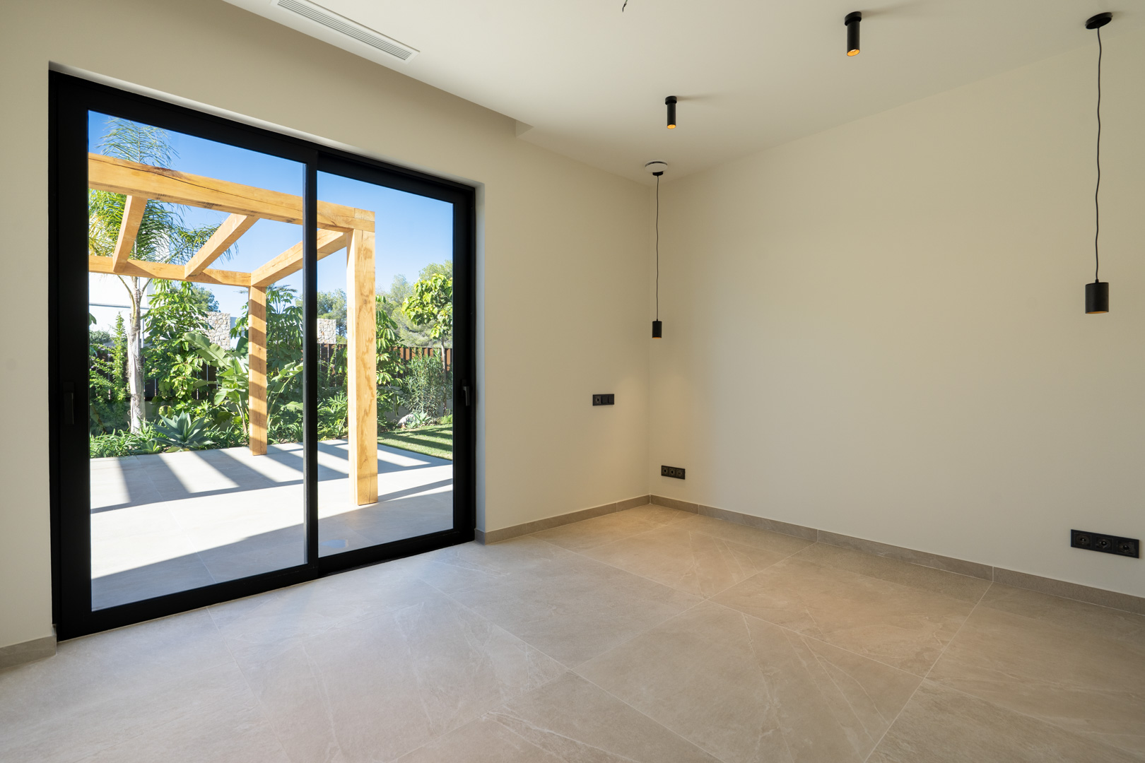 Minimalist interior with ceramic flooring and large sliding doors leading to an outdoor terrace featuring a wooden pergola and Mediterranean landscaping.