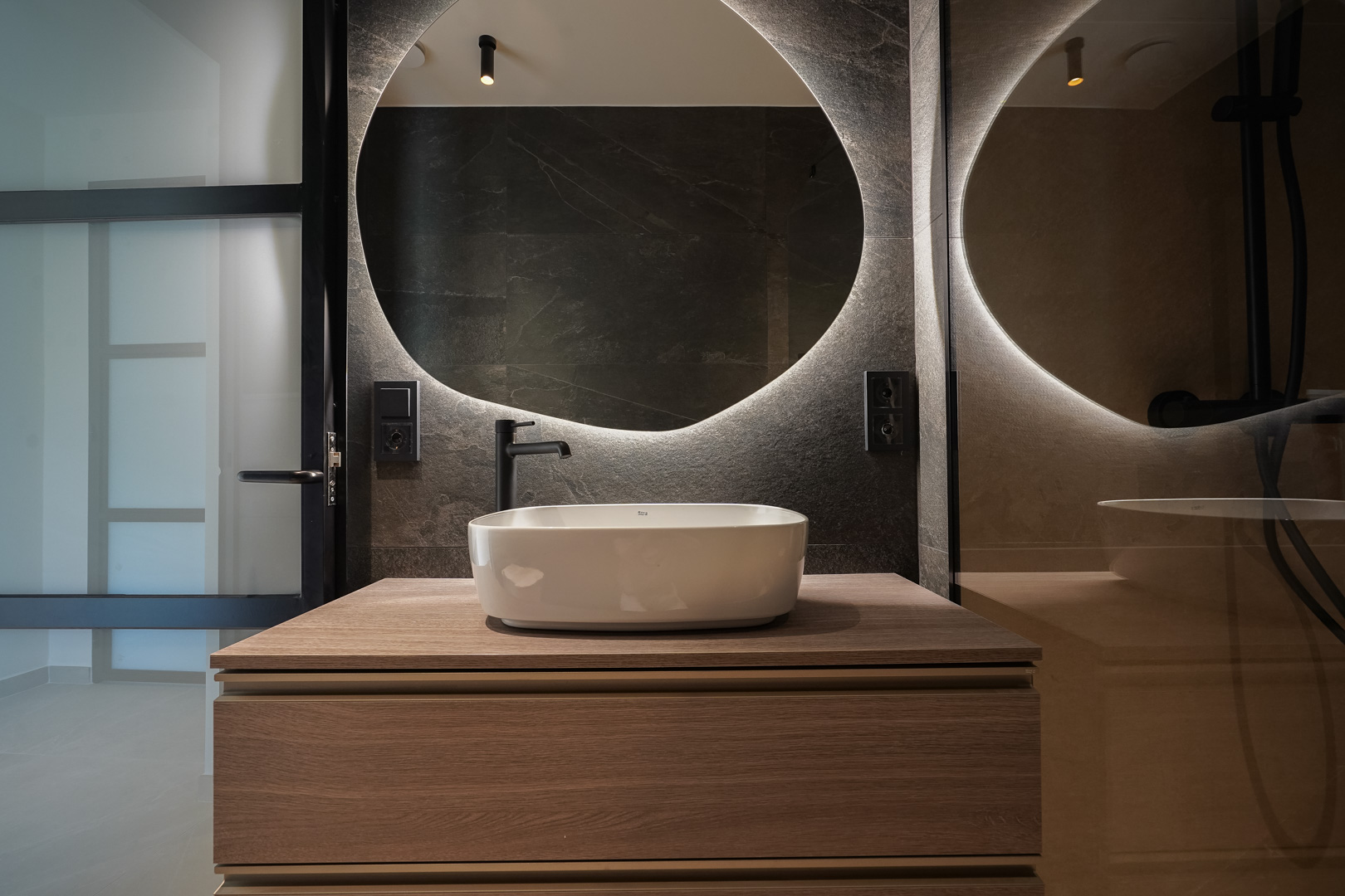 Modern bathroom featuring a white vessel sink on a wooden vanity, a circular backlit mirror, and dark stone-textured walls with matte black fixtures.