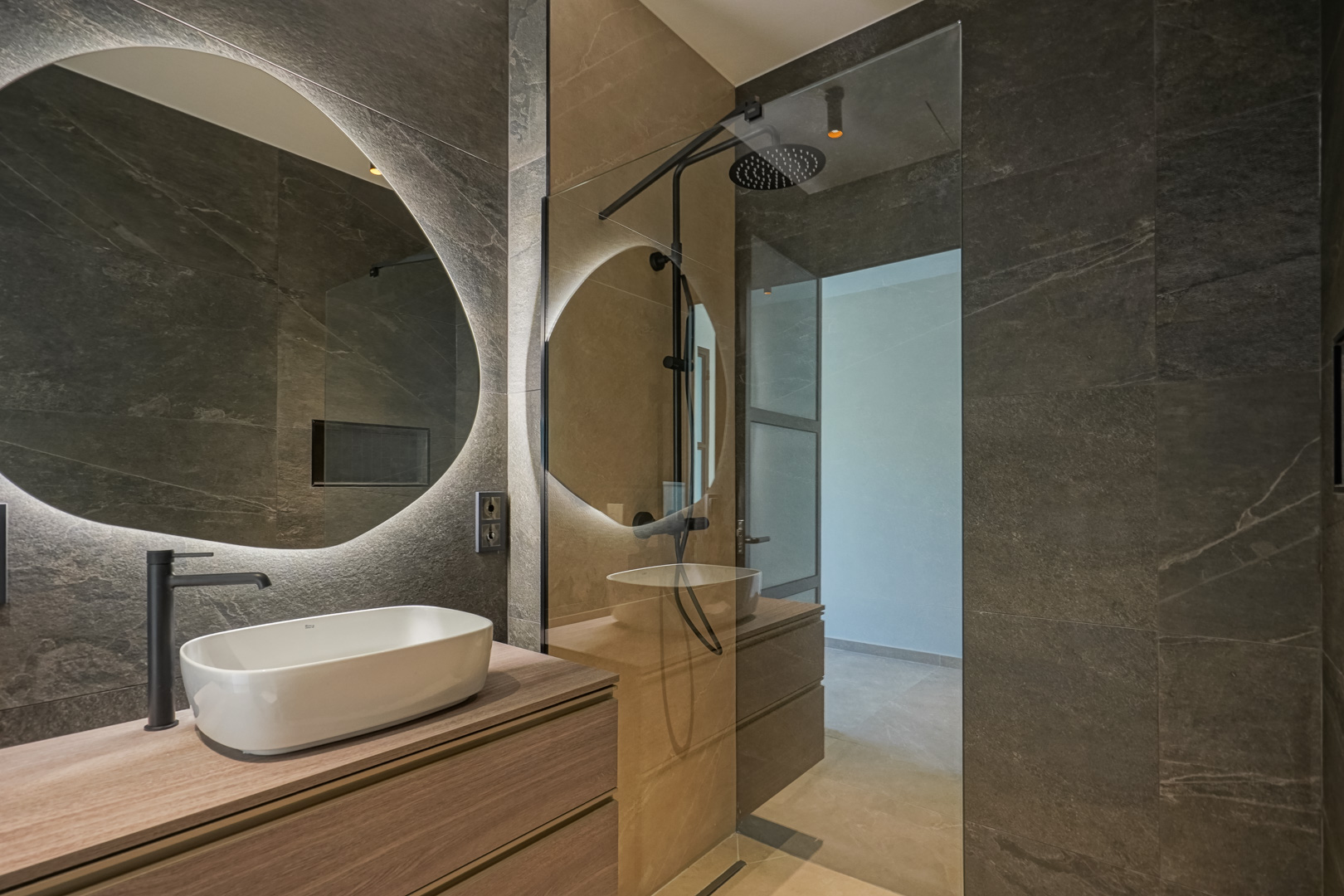 Modern bathroom featuring dark stone-effect tiles, a wooden vanity with a white vessel sink, and a circular backlit mirror next to a walk-in shower.
