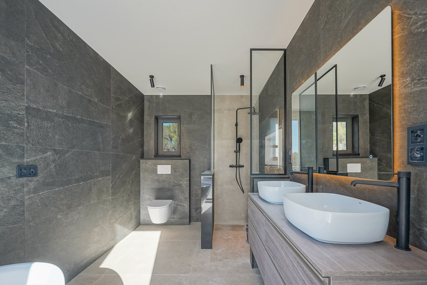 Modern bathroom featuring grey stone-effect tiles, double vessel sinks on a wooden vanity, matte black fixtures, and a walk-in shower with a glass partition.