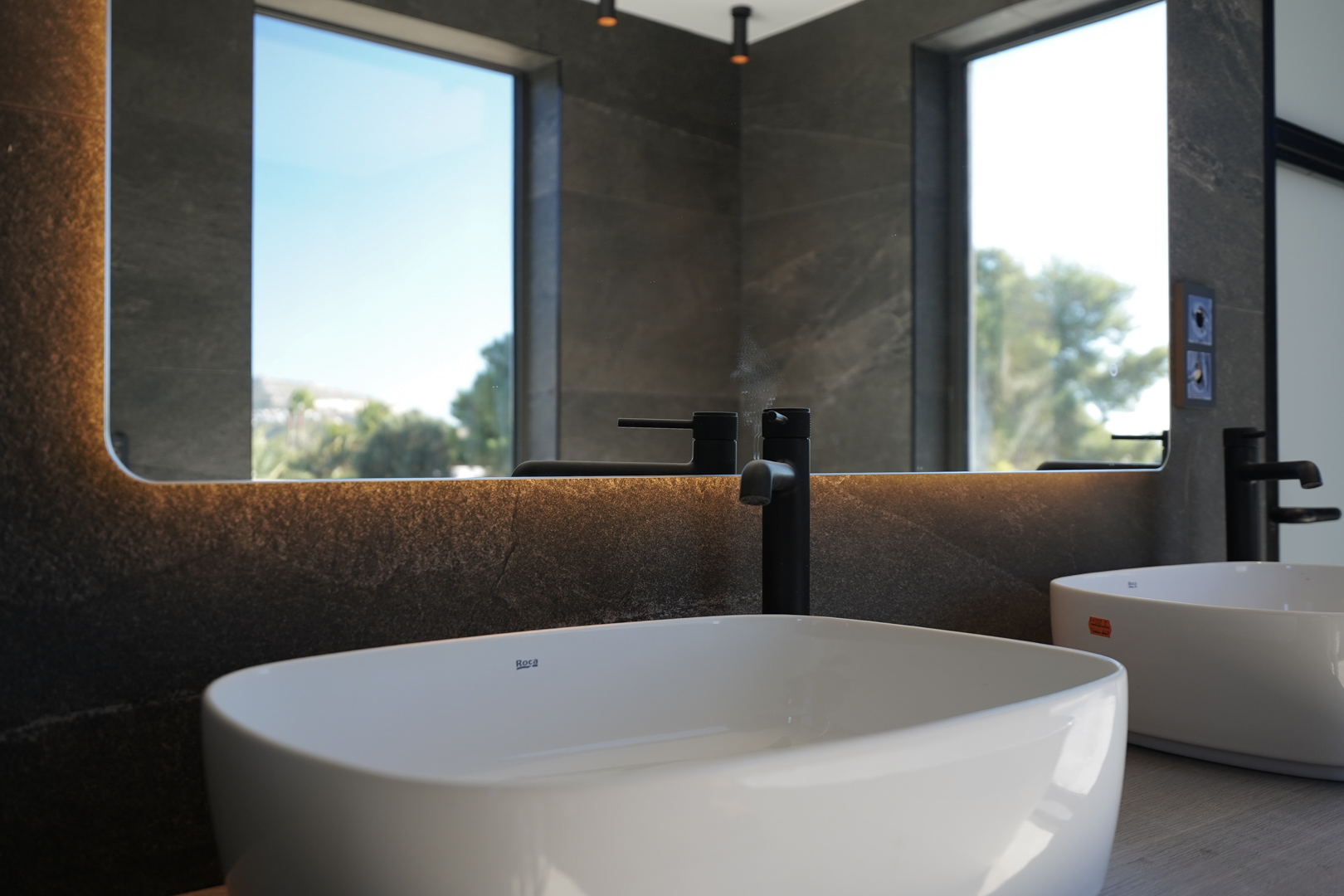 Modern bathroom featuring Roca vessel sinks, matte black faucets, and a backlit mirror against dark stone-effect tiling with natural light.