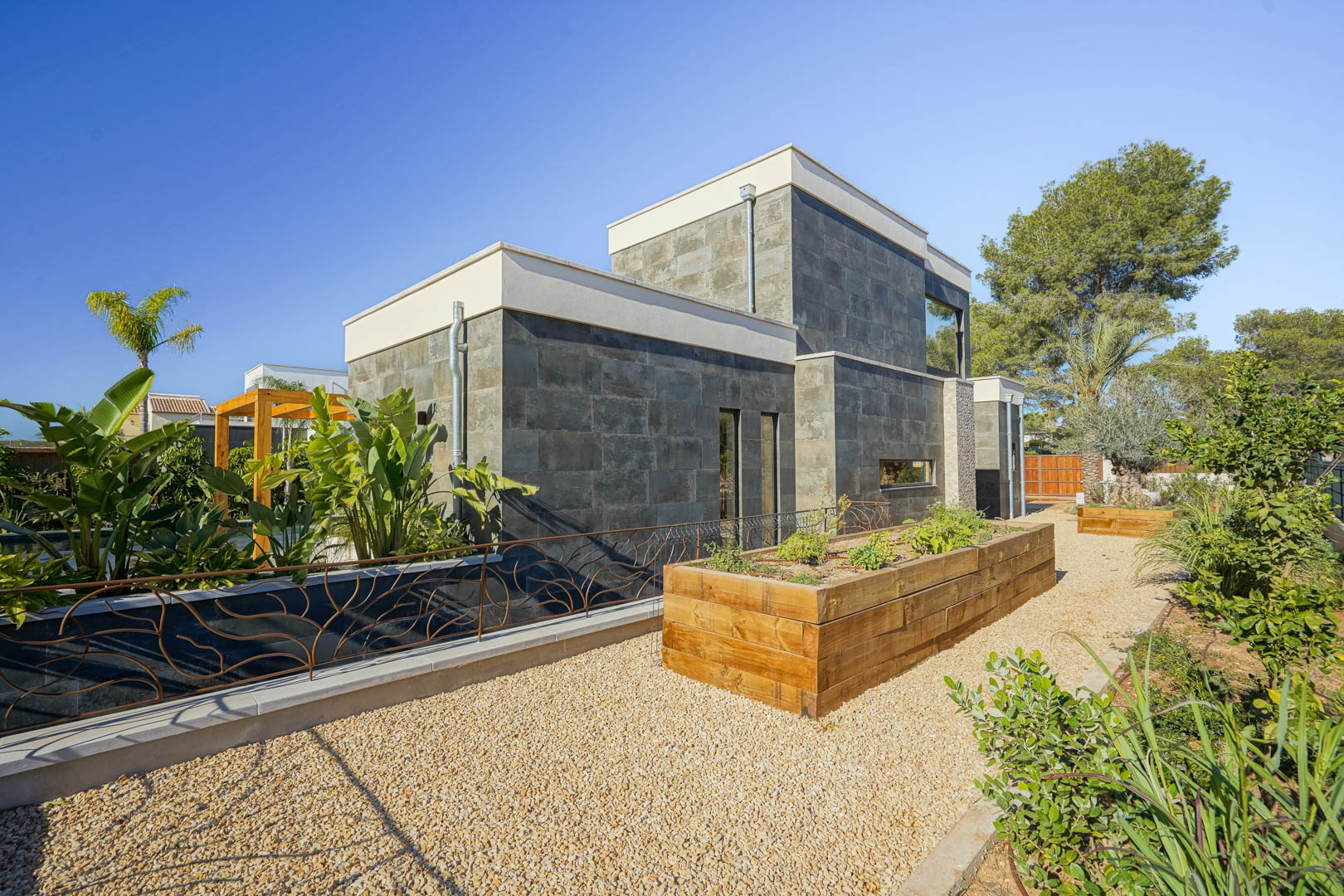 Modern exterior featuring dark stone cladding, raised wooden herb beds, and a gravel path within a landscaped Mediterranean garden.