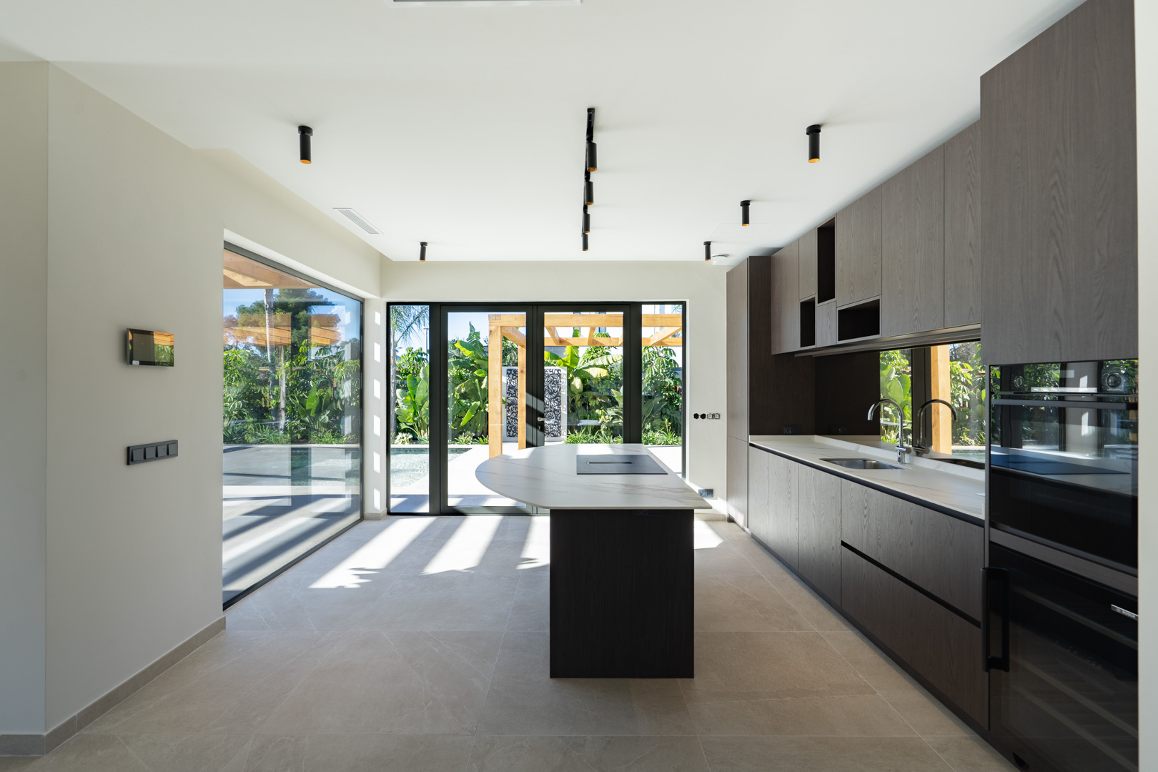 Modern kitchen featuring a central island with stone countertop, dark wood cabinetry, and integrated appliances. Floor-to-ceiling glass doors offer views of the pool and garden.