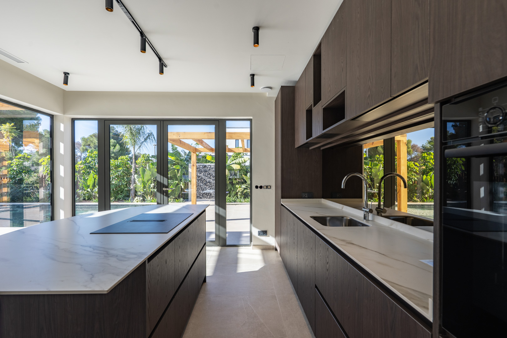 Modern kitchen featuring a large island with marble-effect countertops and dark wood cabinetry. Floor-to-ceiling glass doors open to the pool area and pergola.