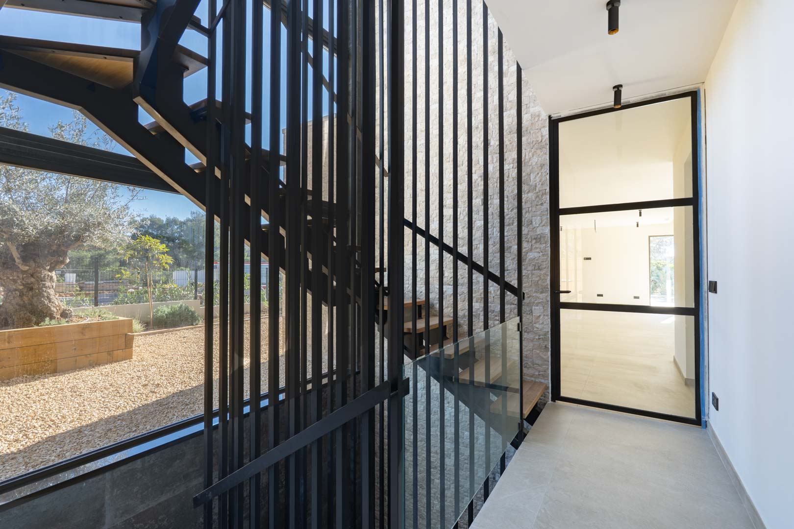 Modern staircase featuring wooden treads, black metal slats, and a stone accent wall, illuminated by natural light from large windows overlooking an olive tree.