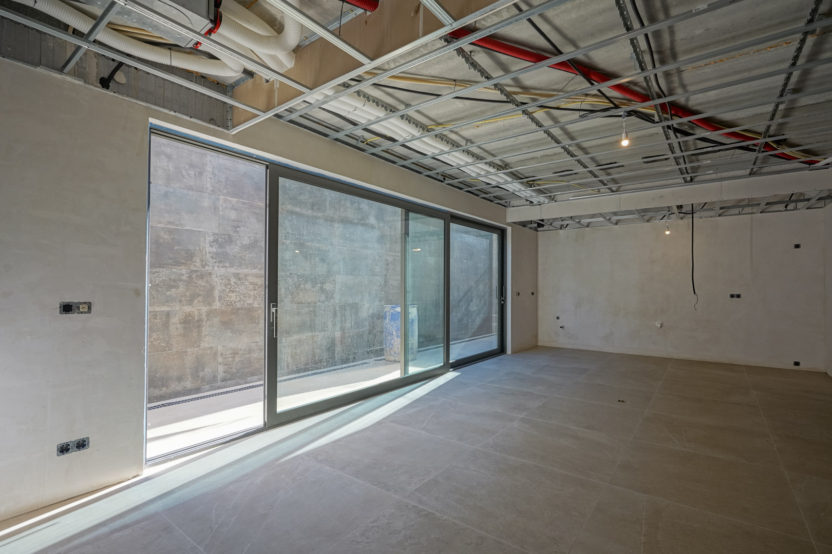 Open-plan basement under construction featuring large sliding glass doors, large-format floor tiles, and exposed ceiling infrastructure for ventilation and electrical systems.