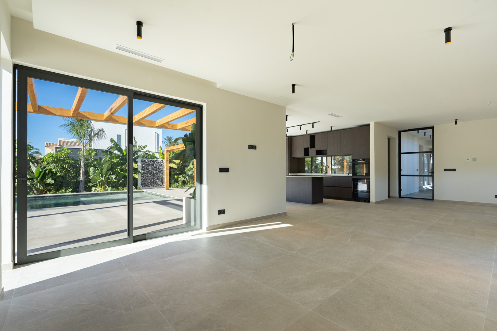 Open-plan living area featuring porcelain flooring and large sliding doors opening to the pool and iroko pergola. A dark wood designer kitchen completes the space.