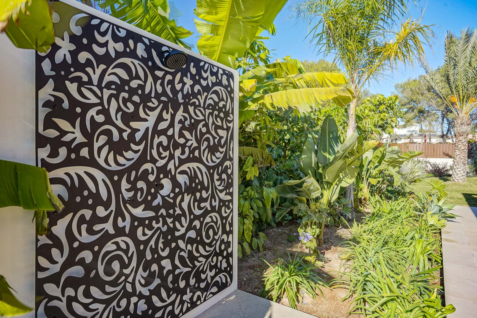 Outdoor shower featuring a decorative laser-cut wall panel and black fixtures, surrounded by lush Mediterranean greenery and banana trees.