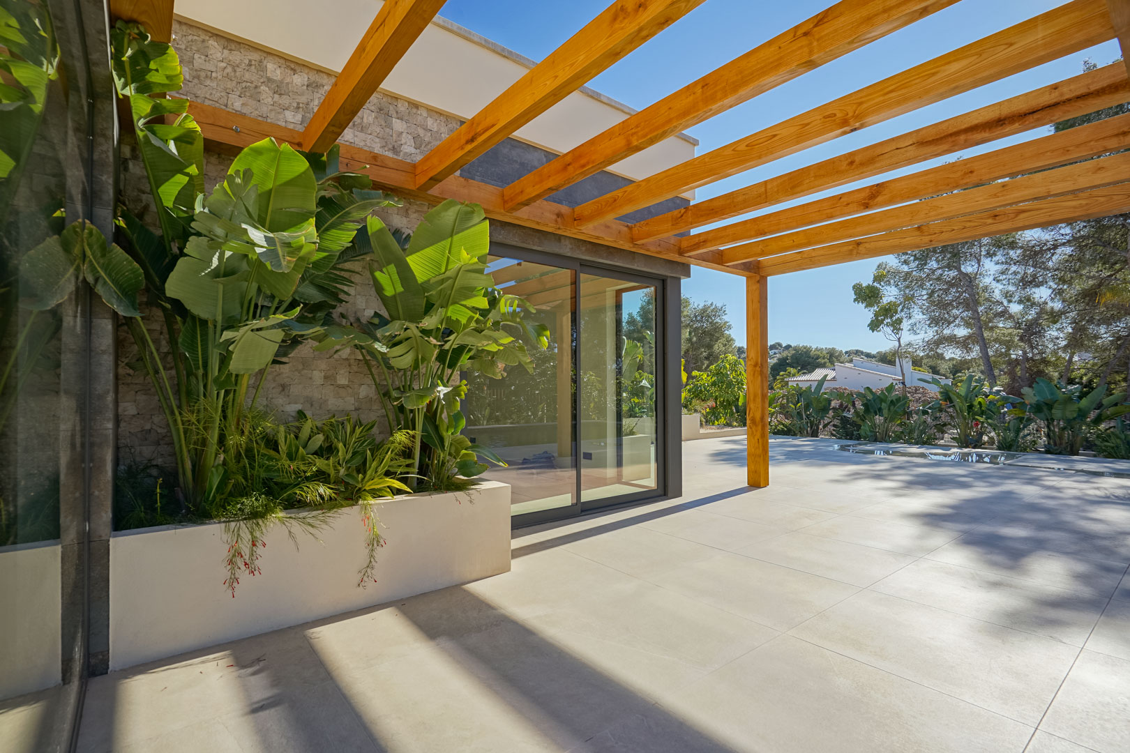 Outdoor terrace featuring an iroko wood pergola, large-format tiling, and a built-in planter with lush tropical greenery against stone-clad walls.