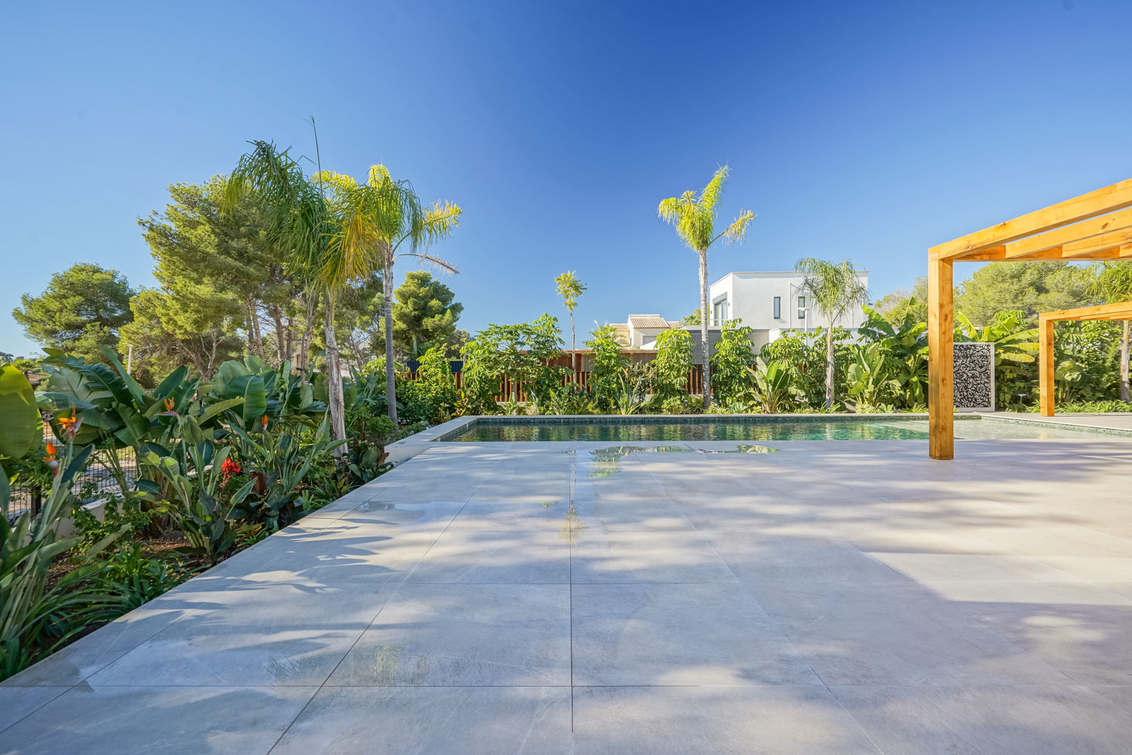 Porcelain tile terrace adjacent to a Bali-style pool, featuring iroko wood pergolas and a lush Mediterranean garden with tropical palms.