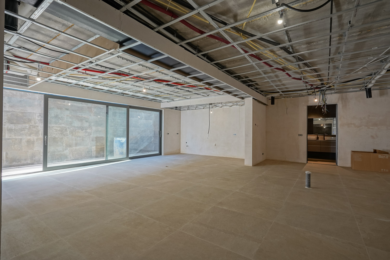 Spacious basement area under construction featuring large-format floor tiles, sliding glass doors for natural light, and exposed ceiling infrastructure.