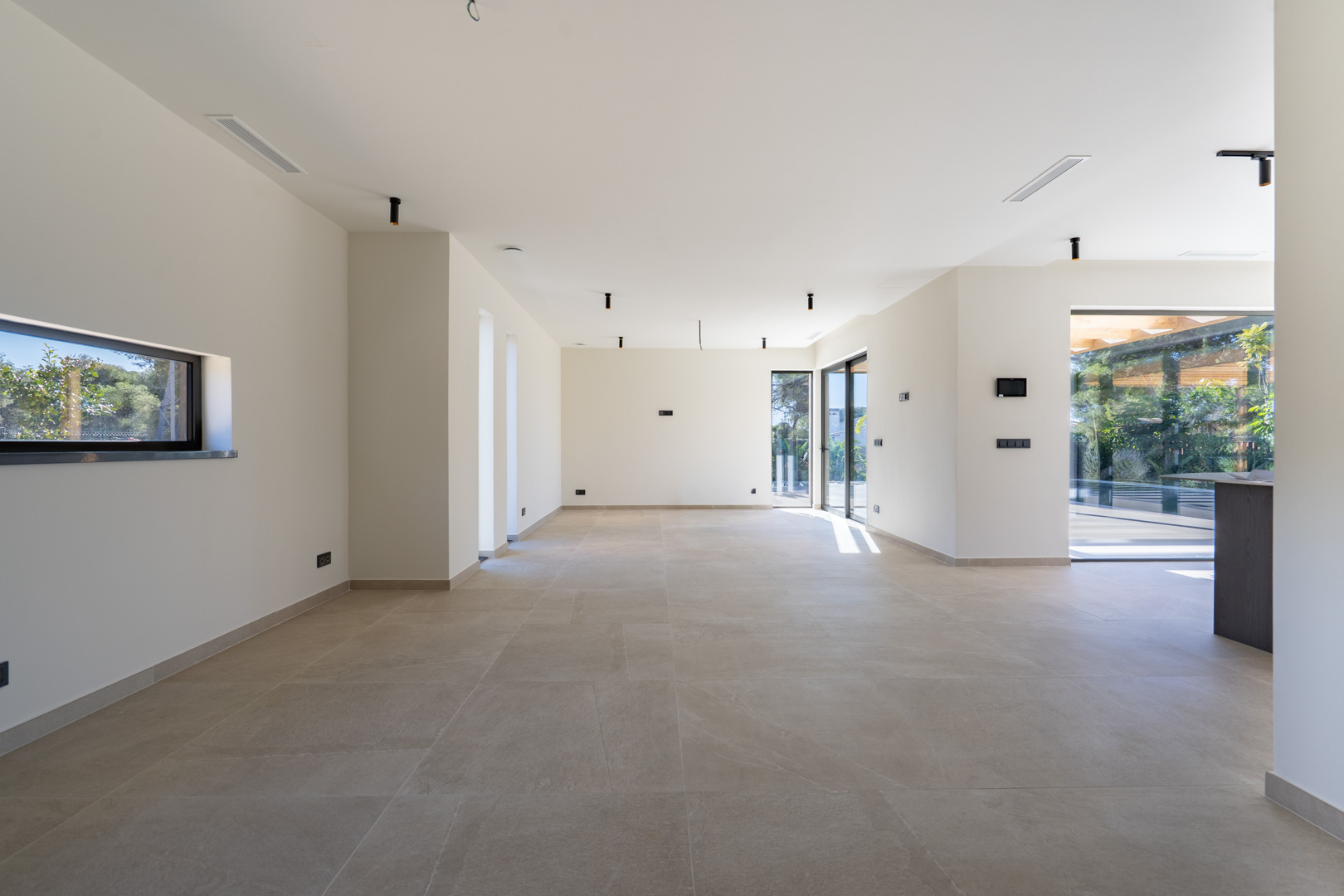 Spacious open-plan living area featuring large-format stone tiles, minimalist track lighting, and floor-to-ceiling glass doors opening to the garden.