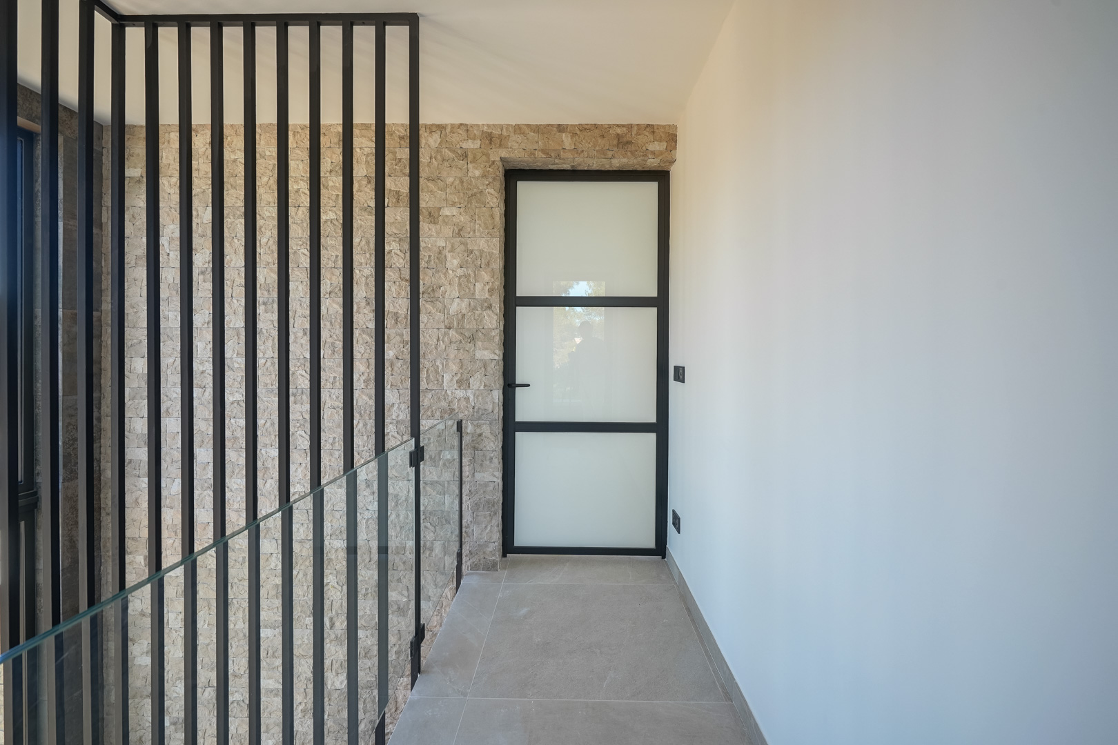 Upper floor landing featuring a natural stone accent wall, glass balustrade with black metal slats, and an industrial-style frosted glass door.
