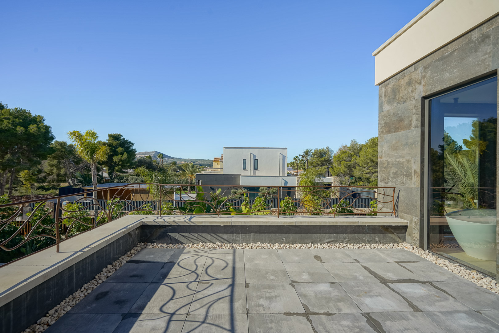 Upper-floor private terrace featuring grey stone tiling, decorative iron railings, and views of the surrounding Mediterranean landscape and neighborhood.