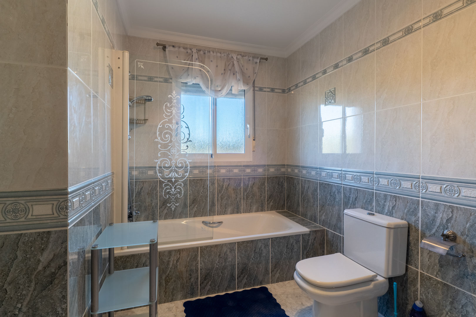 Bathroom featuring a built-in bathtub with an etched glass screen and two-tone tiling. A frosted window provides natural light and ventilation.