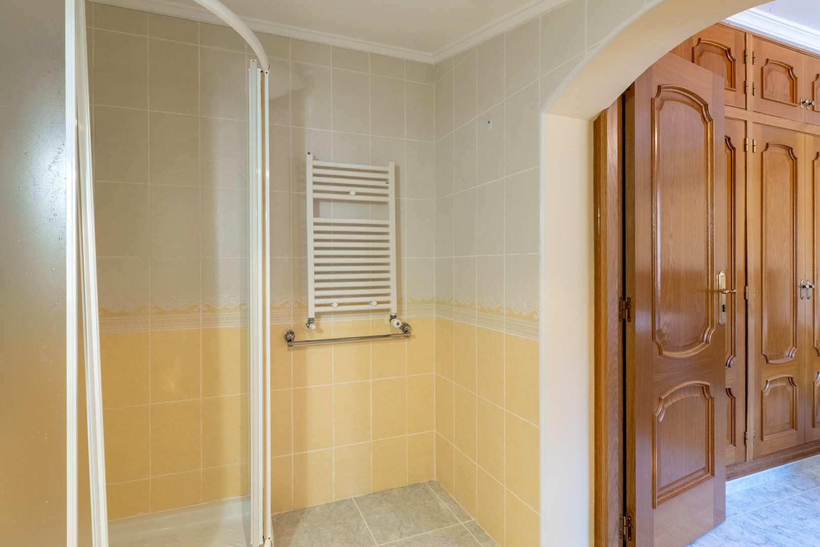 Bathroom featuring two-tone tiling, a towel radiator, and corner shower, connected via an archway to a bedroom with classic built-in wooden wardrobes.