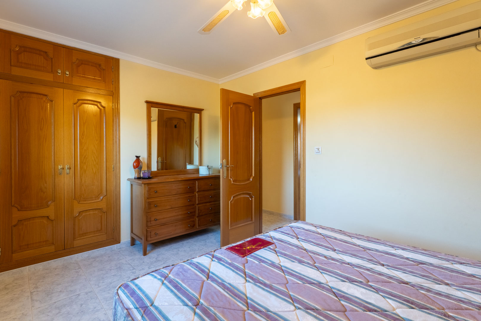 Bedroom featuring built-in wooden wardrobes, a matching dresser with mirror, split-system air conditioning, and a ceiling fan on light tiled flooring.