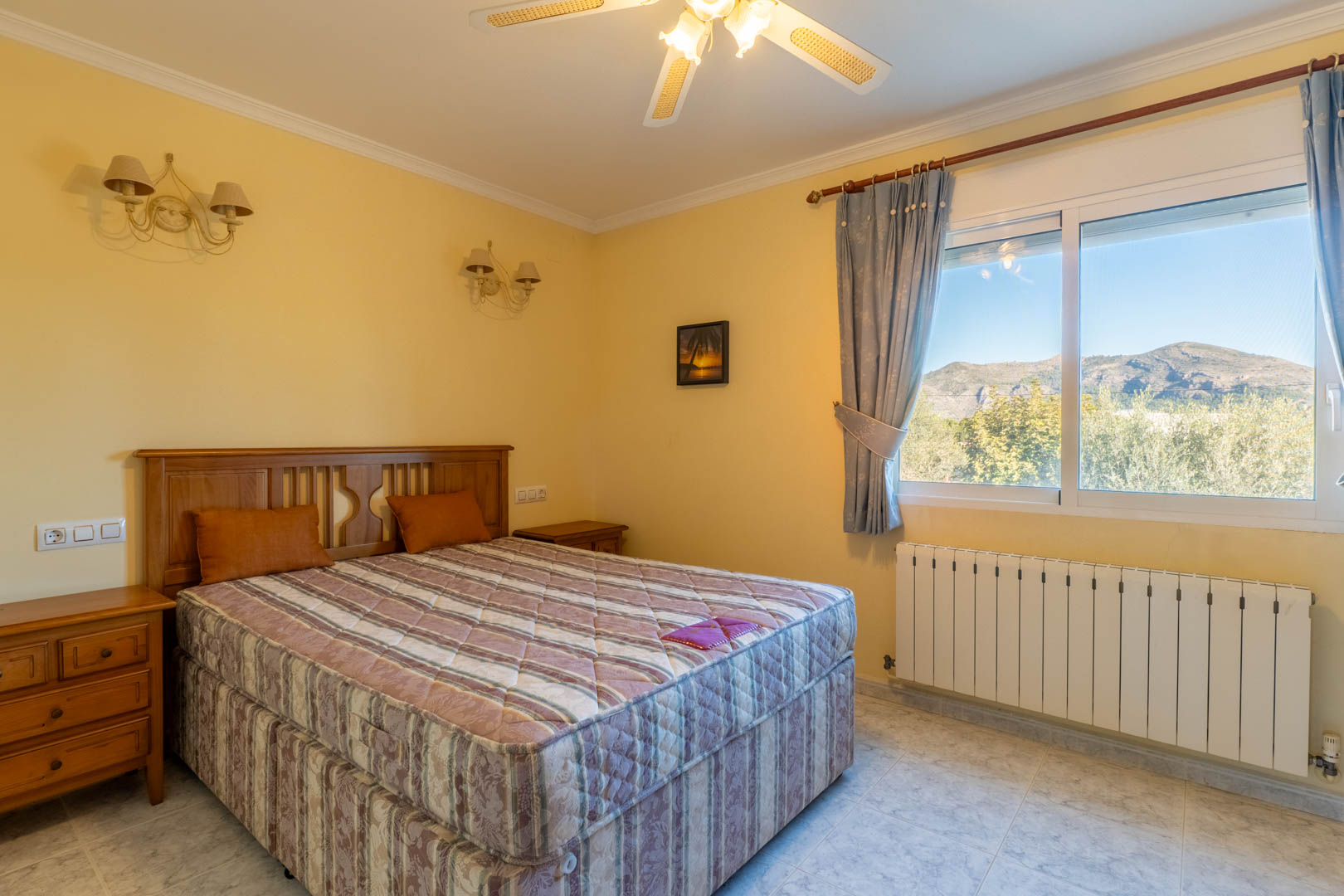Bright bedroom featuring classic wooden furniture, a ceiling fan, and a large window offering scenic mountain views and abundant natural light.
