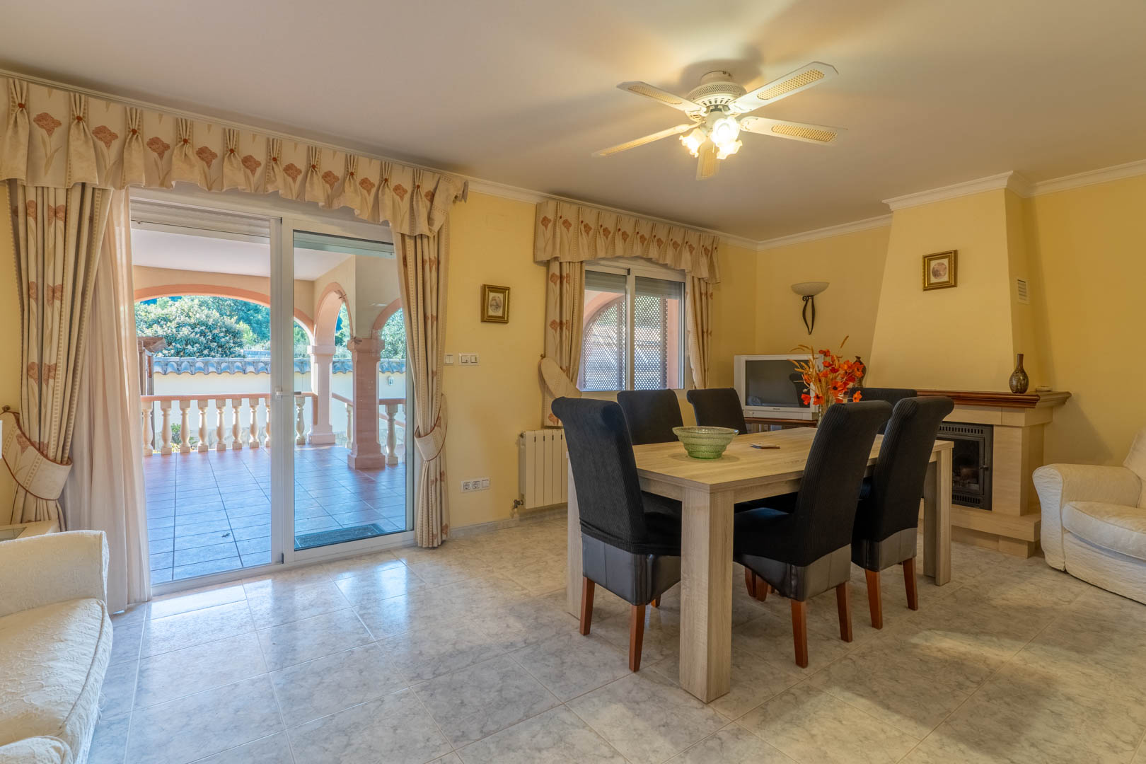 Bright dining area featuring a classic fireplace, wooden table, and direct access to the arched terrace through sliding glass doors.