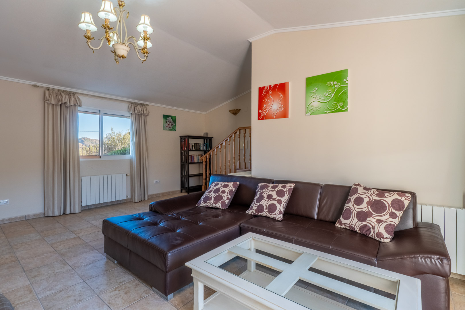 Bright living space with a dark brown leather sectional, white coffee table, and tiled flooring. Features include a wooden staircase, radiator, and classic chandelier.