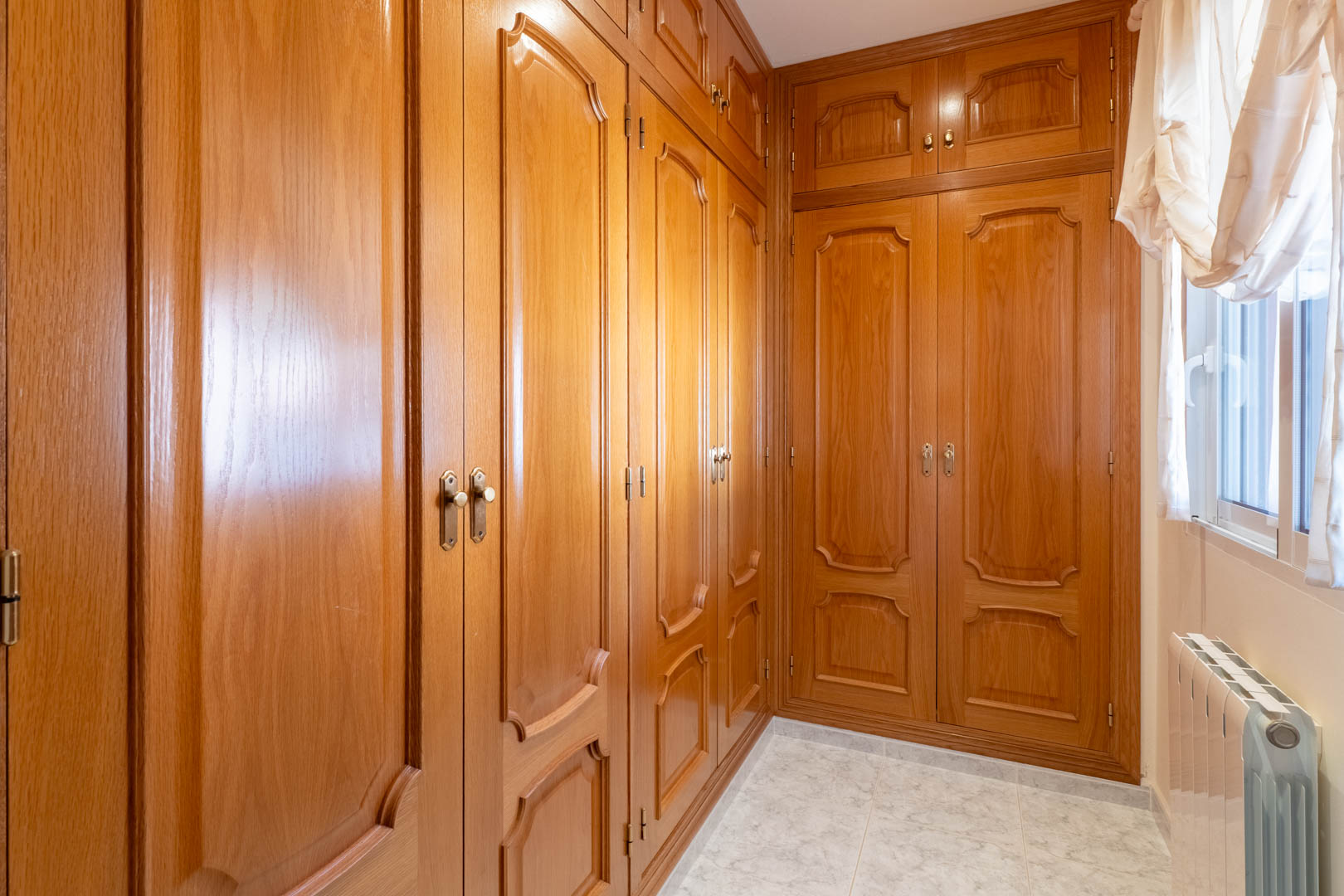 Dressing area featuring extensive built-in wooden wardrobes with classic paneling, ceramic tile flooring, and a window providing natural light.