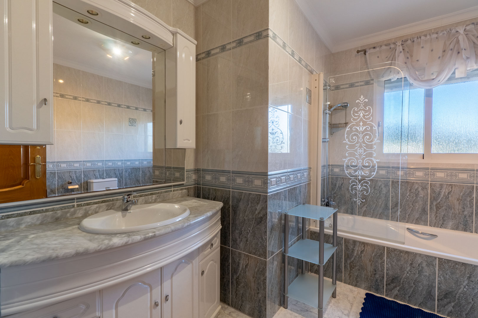 Full bathroom featuring a marble-topped vanity, white cabinetry, and a bathtub with a decorative glass screen. Tiled walls with ornamental borders and natural light.
