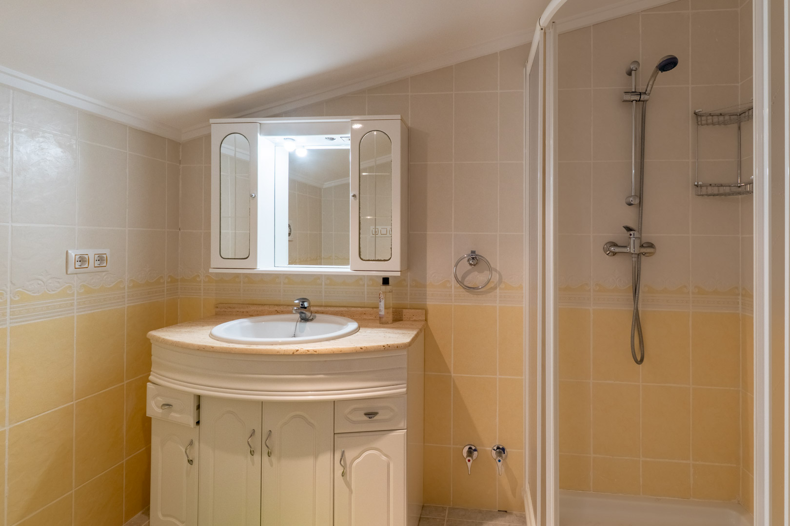 Functional bathroom featuring a marble-topped vanity, white cabinetry, and a walk-in shower, finished with cream-toned tiling and a mirrored wall cabinet.