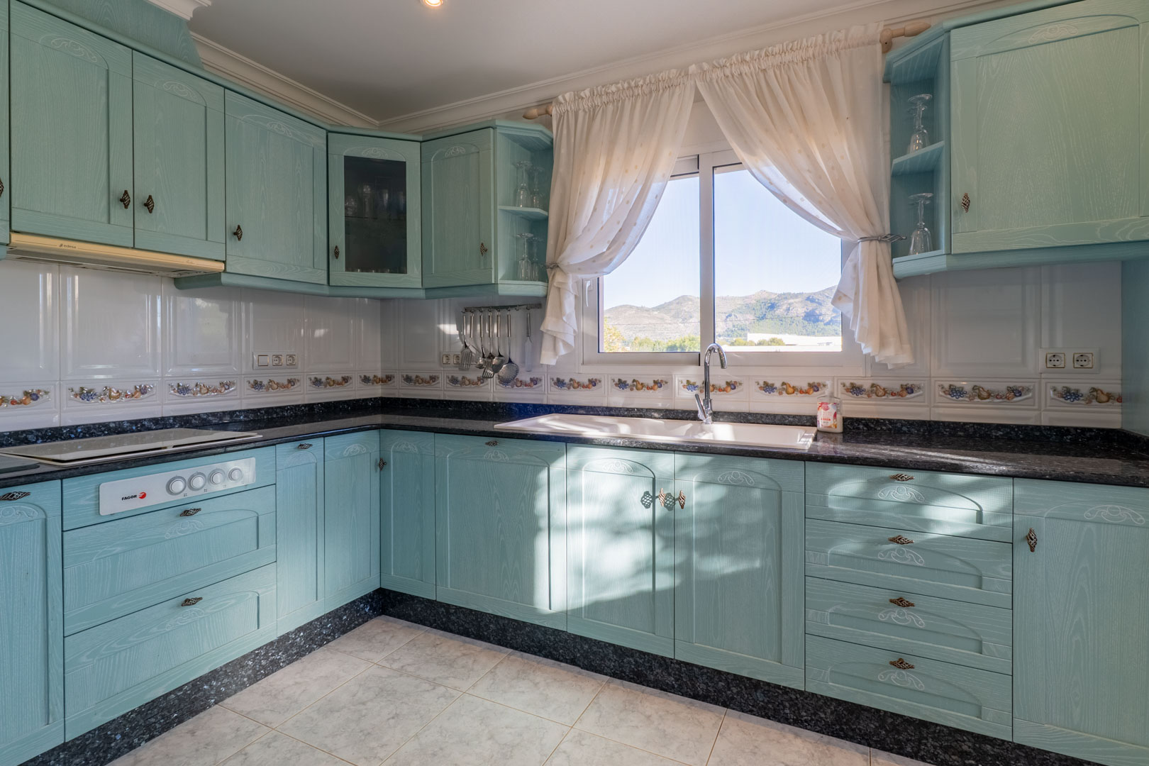 Independent kitchen featuring turquoise cabinetry, dark granite countertops, and a window framing scenic mountain views of the Jalón Valley.