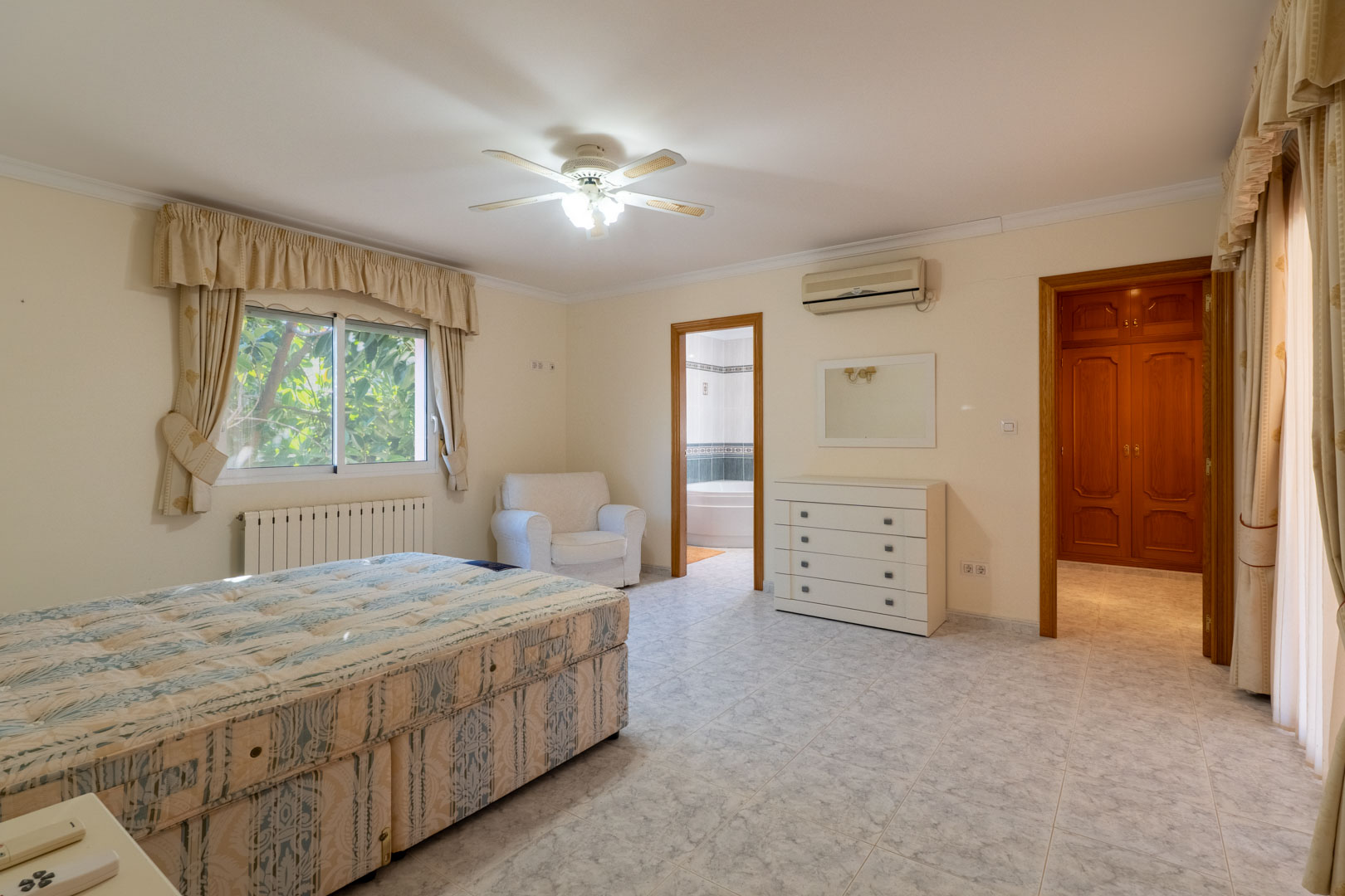 Master bedroom featuring an ensuite bathroom and walk-in closet. Equipped with ceramic flooring, split air conditioning, a ceiling fan, and natural light from large windows.