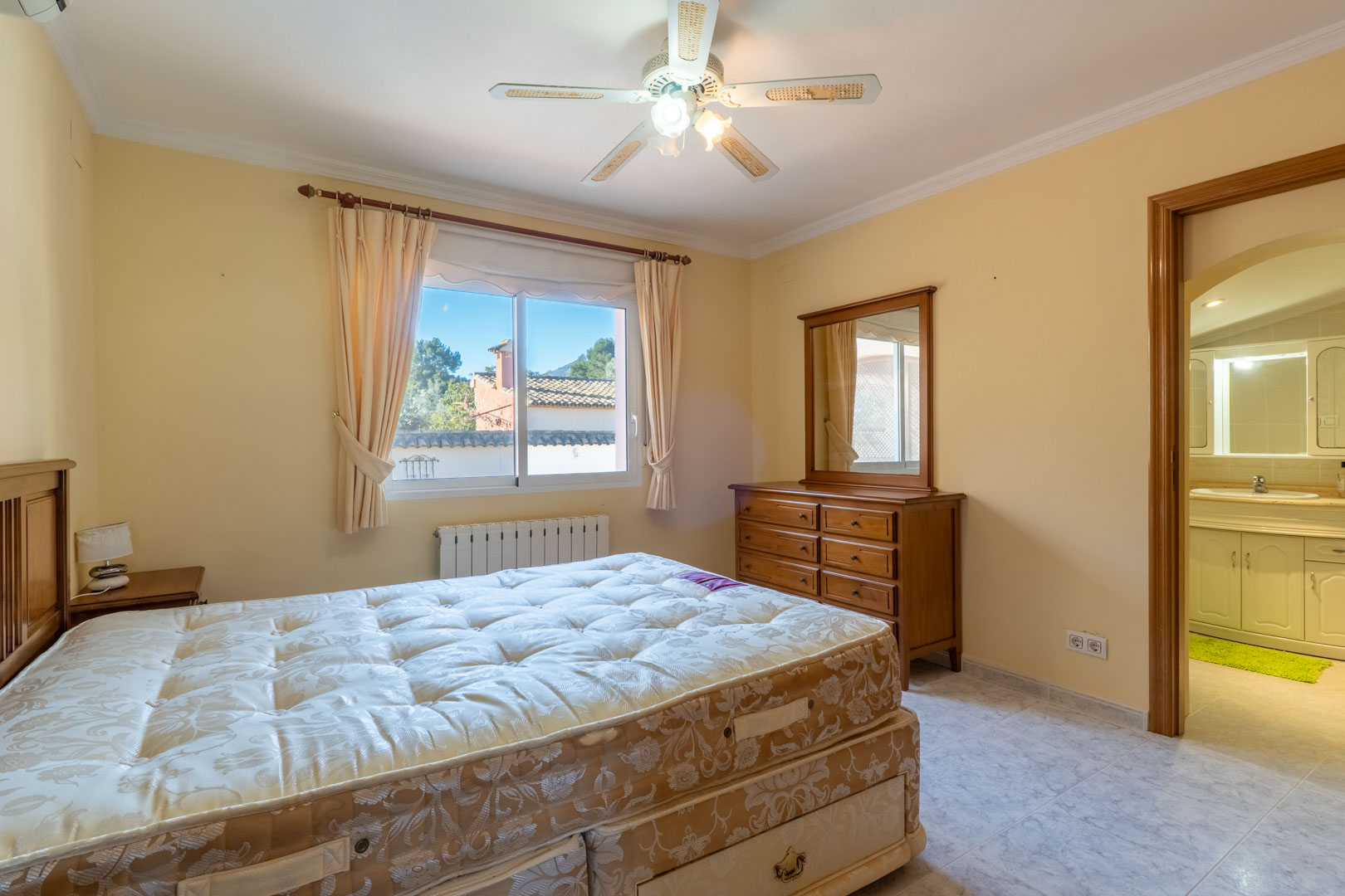 Master bedroom featuring wooden furniture, a ceiling fan, and an en-suite bathroom. The room includes a radiator and a window providing natural light.