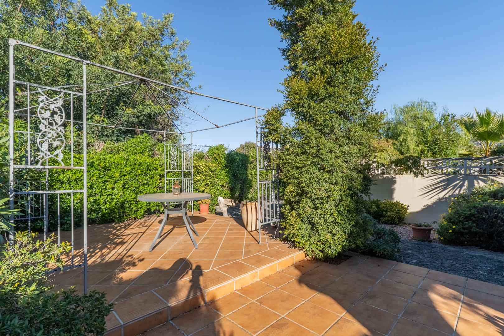 Outdoor seating area featuring a terracotta tile floor, a decorative metal pergola, and a round table, framed by lush Mediterranean greenery and hedges.
