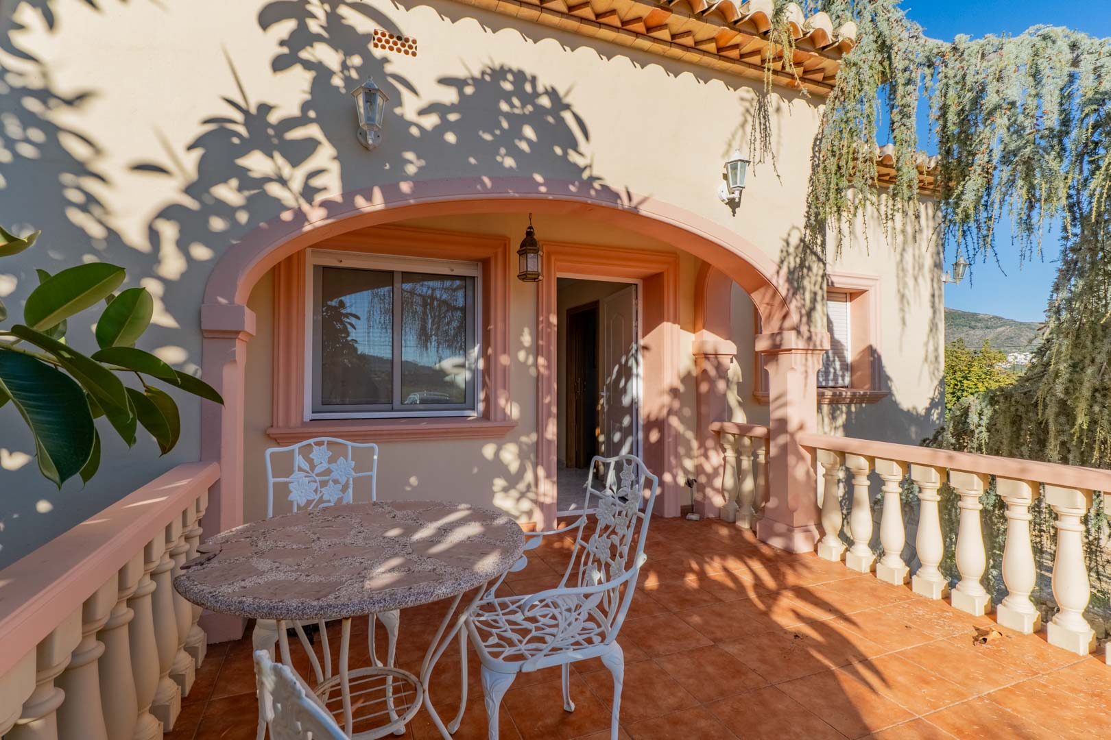 Outdoor terrace featuring Mediterranean arches, terracotta flooring, and white wrought iron furniture, highlighted by natural dappled sunlight.