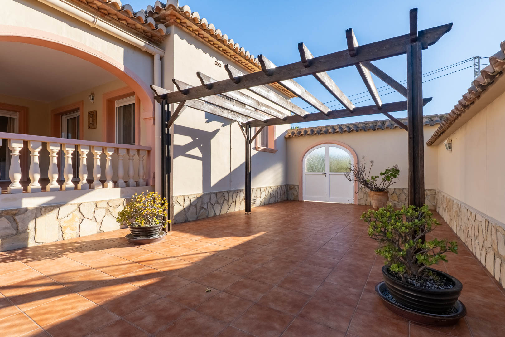 Outdoor terrace featuring terracotta tiling, a wooden pergola, and stone-clad walls. The space connects to a raised porch with a classic balustrade.