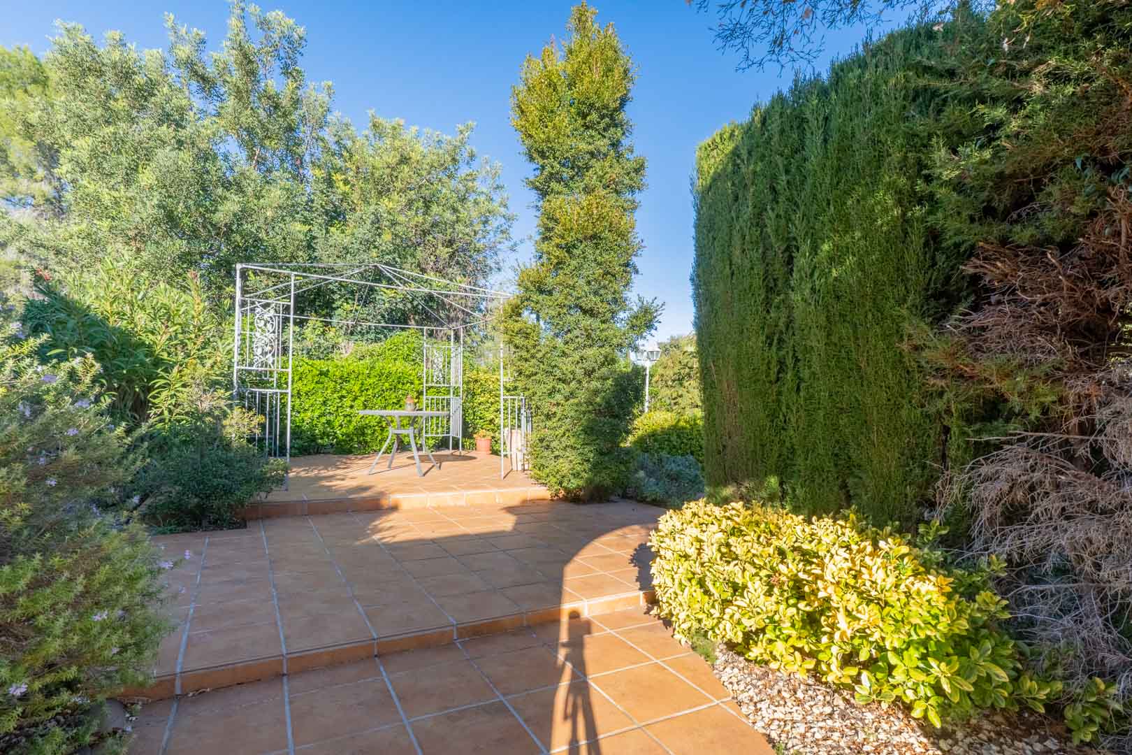 Outdoor tiled terrace featuring a white metal gazebo and seating area, surrounded by mature Mediterranean hedges and lush green landscaping.