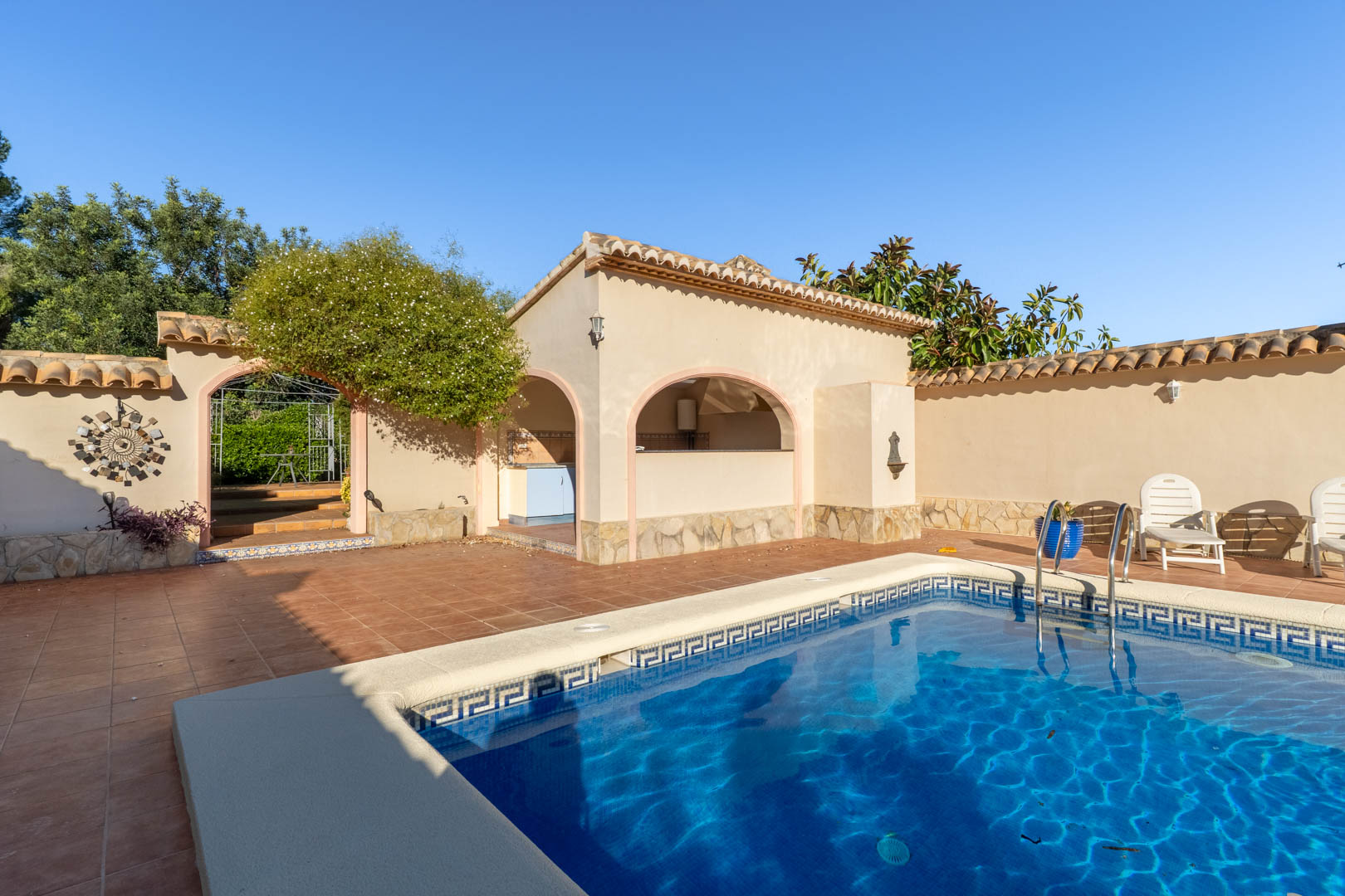 Pool area featuring terracotta tiling, a covered outdoor kitchen with arched openings, and stone-clad walls under a clear sky.