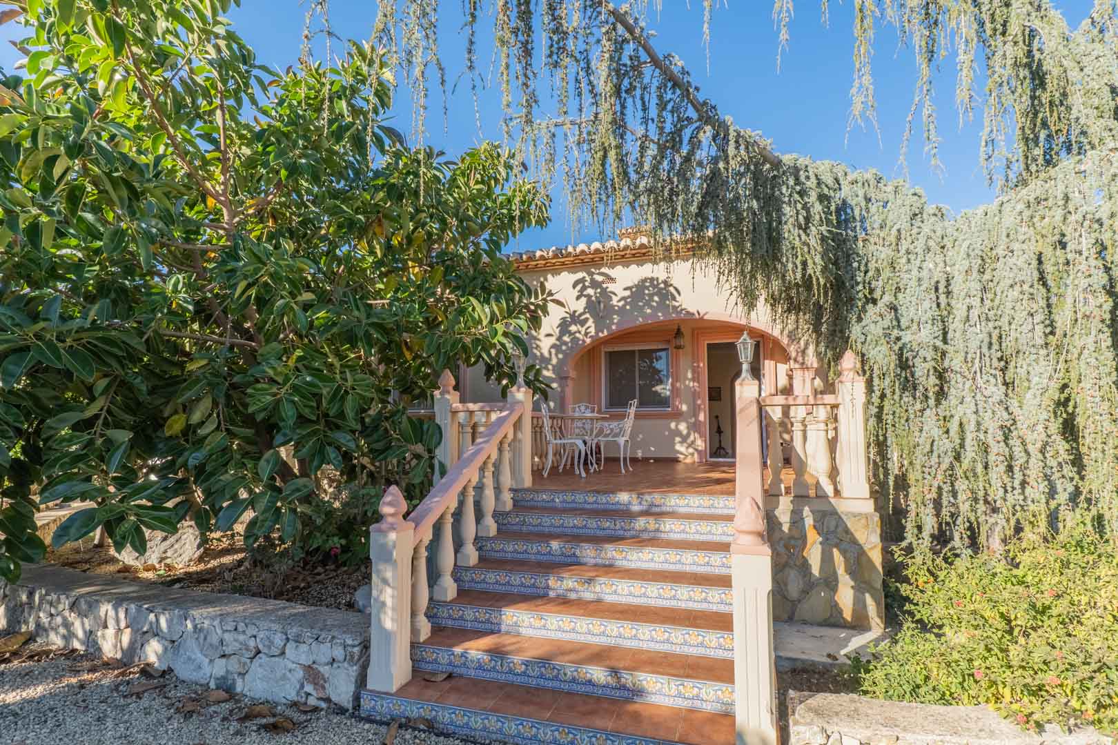 Tiled exterior stairs with decorative risers lead to an arched terrace featuring white metal furniture, framed by lush Mediterranean trees and a stone base.