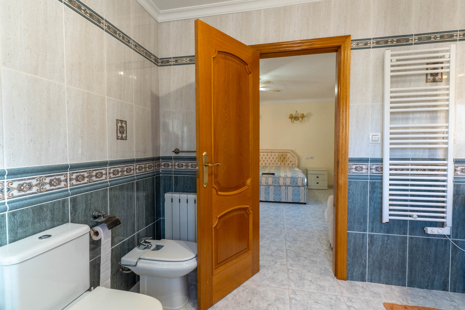 View from the en-suite bathroom into the master bedroom, featuring decorative ceramic tiling, a white towel radiator, and solid wood doors.