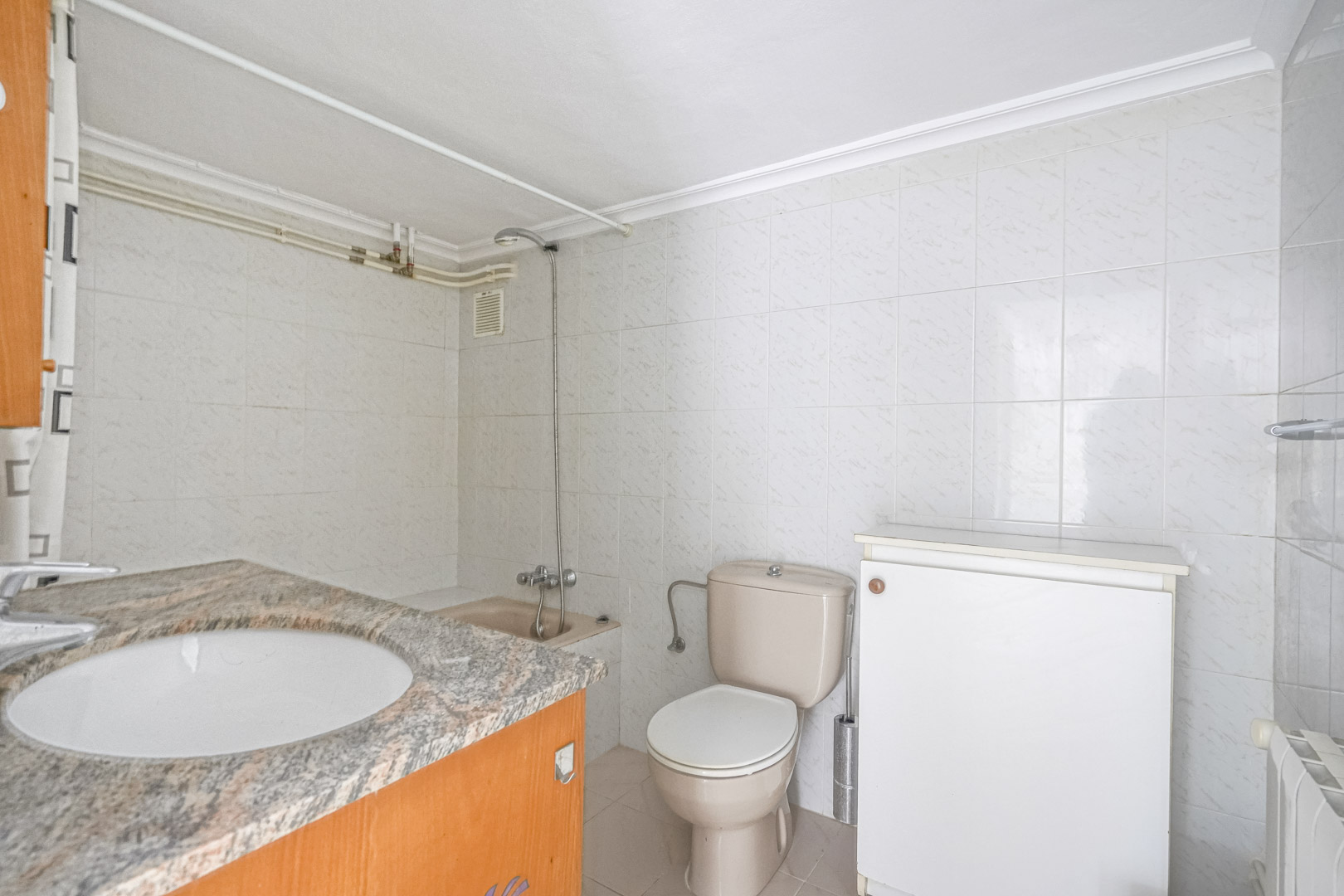 Bathroom featuring a granite countertop with an inset sink, beige toilet, and white tiled walls. Includes a shower area with exposed piping and a wall-mounted radiator.