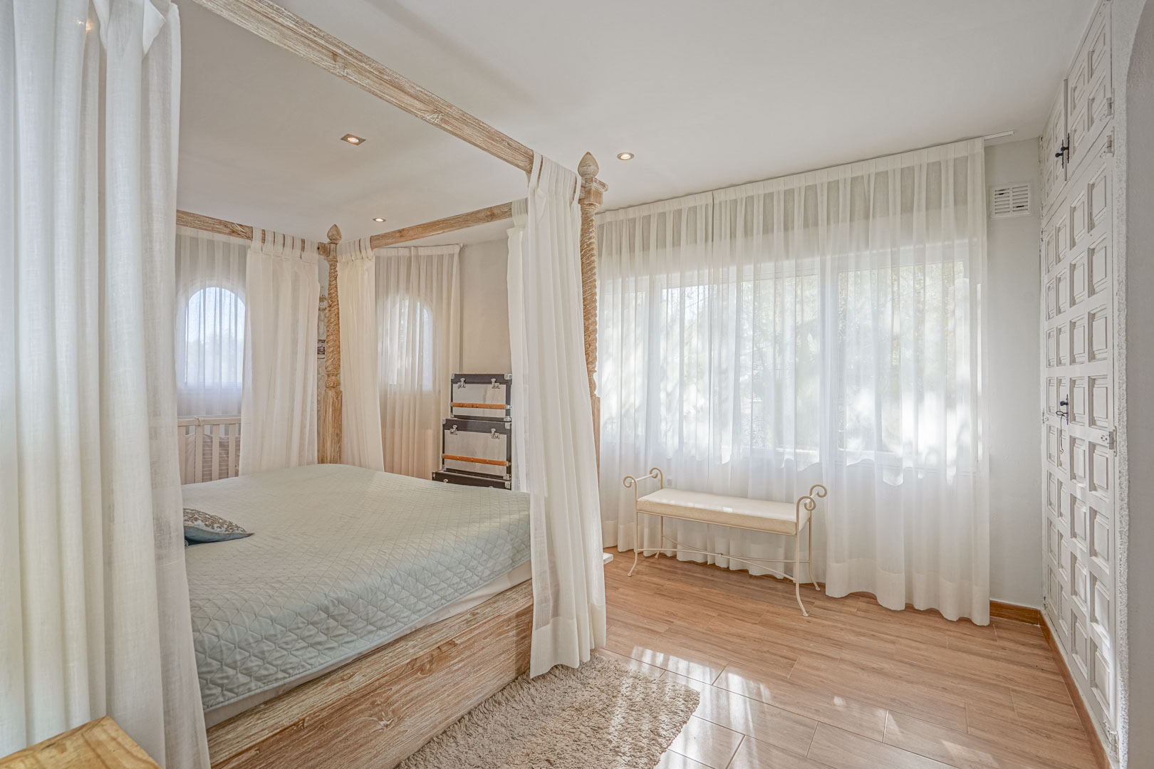 Bedroom featuring a light wood four-poster bed, wood-effect ceramic flooring, and large windows with sheer curtains that filter soft natural light.