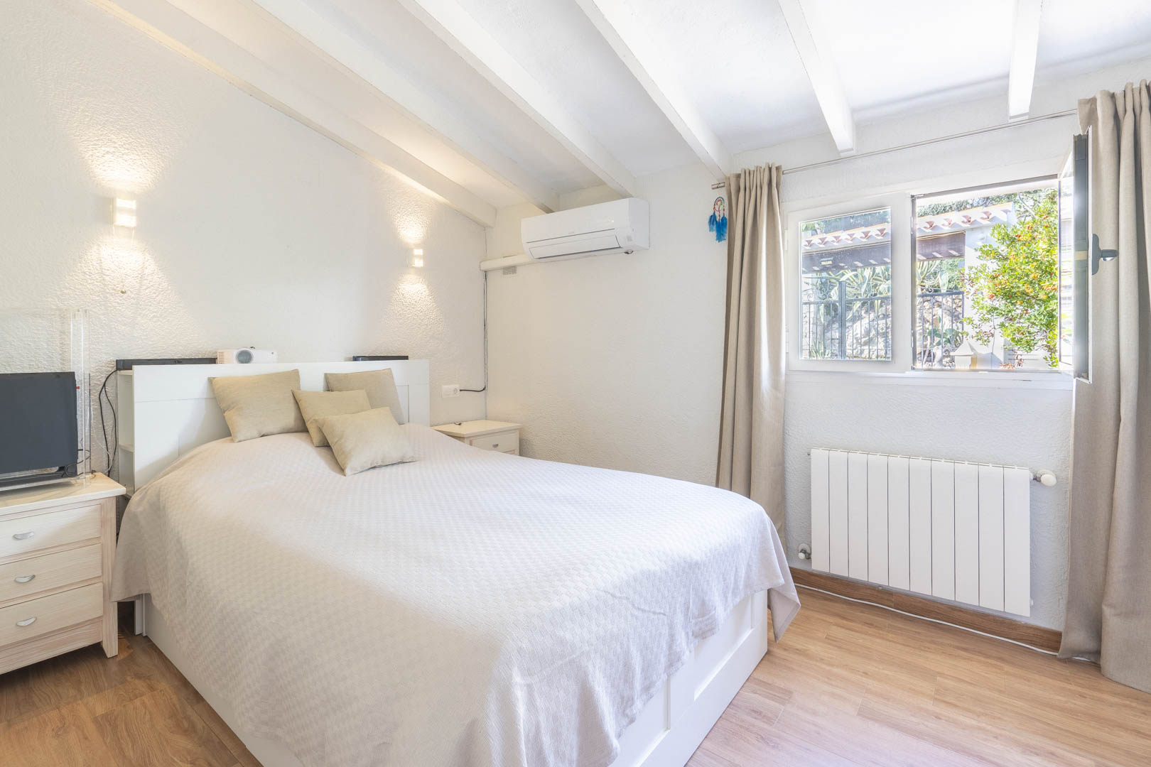 Bedroom featuring white exposed ceiling beams, split air conditioning, and a wall radiator. Includes laminate wood flooring and natural light.