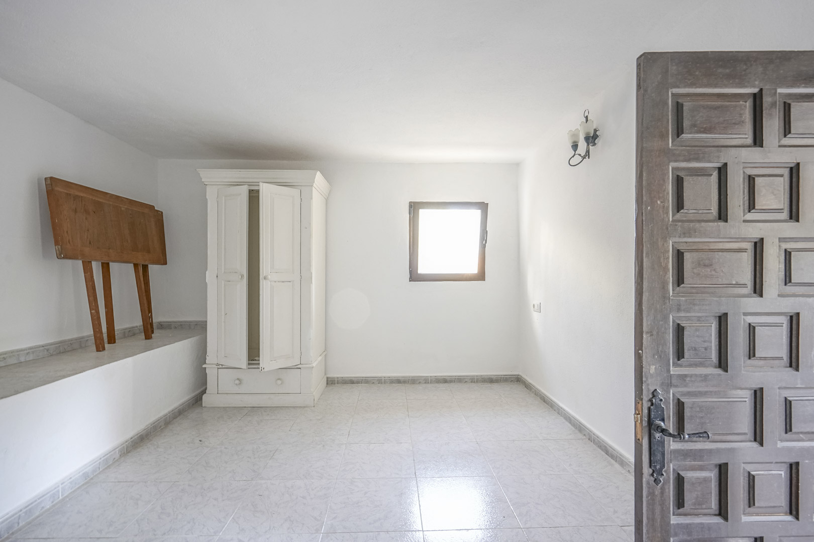 Bright room featuring light-colored tiled flooring, white walls, and a traditional carved wooden door. A freestanding white wardrobe and a small window provide natural light.